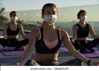 Fit sporty young woman wear medical face mask for safety meditating keeping social distance with female friends practicing yoga fitness exercise together on sunrise at retreat group class outdoors. - Powered by Shutterstock