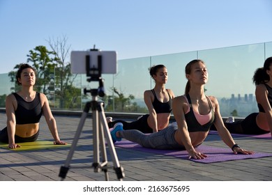 Fit Sporty Young Women Doing Yoga Surya Namaskar Pose Live Stream Online Social Media Training Record Video Blog Remote Group Fitness Tutorial Vlog Workout Class Outdoor On Phone Camera On Tripod.