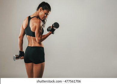 Fit Strong Young Woman Lifting Weights Working Out With Dumbbells Standing With Her Back To The Camera Flexing Her Arm On Grey With Copy Space