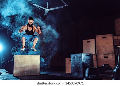 Fit tattoed bearded man jumping onto a box as part of exercise routine. Man doing box jump in the gym. Athlete is performing box jumps.