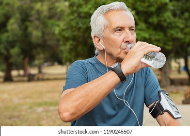 Fit Thirsty Senior Man Drinking Water Before Running. Active Old Man Having A Break During His Jog Routine Outside. Aged Man Drinking Water While Doing Fitness In A Park.