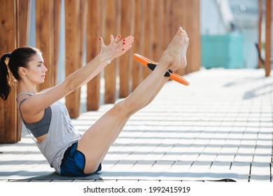 Fit woman doing hollow hold with a pilates hoop between her legs. Outdoors under a wooden frame. Sitting on the pavement. On a sunny day. Striped shadow. - Shutterstock ID 1992415919