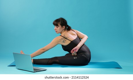 Fit Woman Doing Stretching Exercise And Watching Meditation Video On Laptop In Studio. Athlete Doing Arms And Legs Stretch After Workout Exercise, Following Online Yoga Lesson On Computer.
