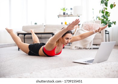 Fit woman doing superman's exercise and watching online workout tutorial on laptop, training in living room