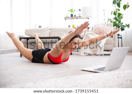 Fit woman doing superman's exercise and watching online workout tutorial on laptop, training in living room