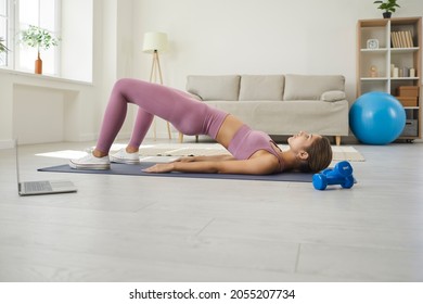 Fit woman doing workout at home. Young lady watching online video lesson on notebook PC, having virtual class with remote instructor, doing strengthening glute bridge gymnastic exercise on fitness mat