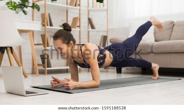 Fit woman doing yoga plank and\
watching online tutorials on laptop, training in living\
room