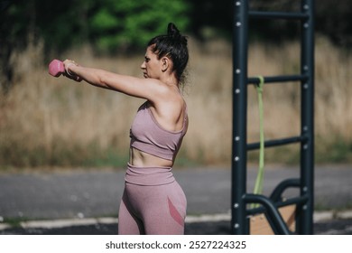 Fit woman engaged in calisthenics workout routine holding pink dumbbells in an outdoor park setting. - Powered by Shutterstock