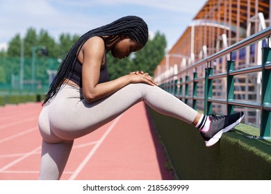 Fit woman with long dark braids stretches legs putting foot on fencing of athletic field in city on sunny summer day. African American female in sportswear tries hard to prepare for upcoming - Powered by Shutterstock