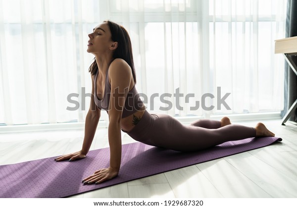 Fit woman making cobra pose on yoga mat,\
exercising in studio over panoramic\
window