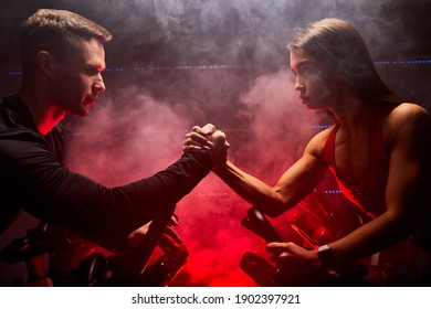 fit woman and man training on smart stationary bike indoors, sport competition in red smoky space. arm wrestling