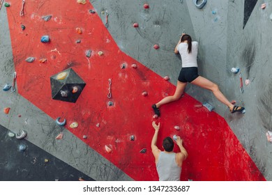 Fit Woman And Man Training At Rock Climbing Wall. Low Angle View. Red, Grey And Black Background.