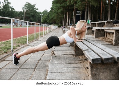 A fit woman performs push-ups on a wooden bench outdoors near a track, focusing on strength and fitness. The surrounding area includes trees and a sports field - Powered by Shutterstock