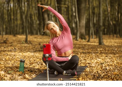 Fit woman in pink top and black leggings demonstrating side stretch during yoga routine while filming with smartphone in an autumn forest. - Powered by Shutterstock