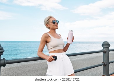 Fit woman relaxes and drinking water leaning on handrail along seaside promenade after running on a road by the sea. Fitness, sport and healthy lifestyle concept.