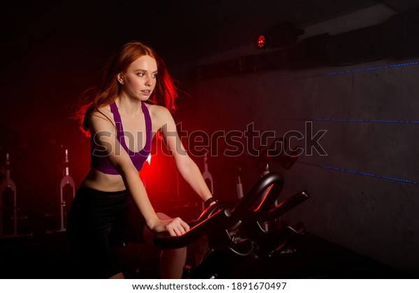 fit\
woman riding on the spinning bike at gym, wearing sportive outfit,\
exercising alone, in smoky room with red neon\
lights