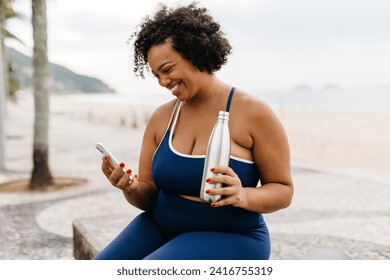 Fit woman in sportswear sitting on a beach promenade, checking her fitness progress on her phone and smiling. Woman engaging in a beach workout and enjoying a healthy lifestyle outdoors near the sea.