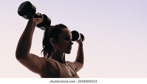 Fit woman in sportswear trains her upper body in a studio, lifting weights and using dumbbells. Her silhouette showcases her toned physique and dedication to her fitness routine. - Powered by Shutterstock