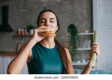 Fit Woman In Sportwear Eating Burger In The Kitchen After Workout. Weight Loss Concept. Food Concept.