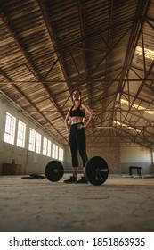 Fit woman standing at gym with barbell on floor at old warehouse. Strong and muscular female resting after training session at workout space in abandoned factory.