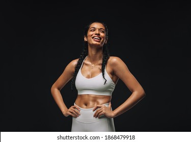 Fit woman standing with hands on waist against a black background. Smiling woman in white sportswear taking break from workout.