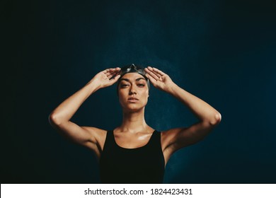 Fit Woman In Swimwear With Swimming Goggles Looking At Camera. Professional Female Swimmer On Dark Background.