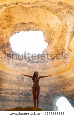 fit woman tourist in bikini standing with spread arms rough in Benagil cave with hole while enjoying summer vacation in tropical country