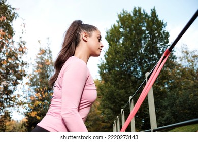 A Fit Woman Is Using A Resistance Band Attached To A Banister. She Is In A London Park In Autumn.