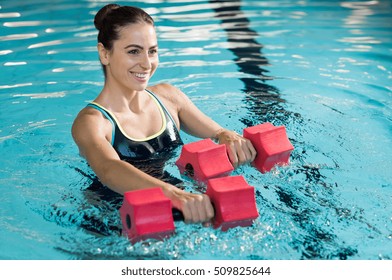 Fit woman working out with foam dumbbell in swimming pool. Woman engaged in doing aqua aerobics in water. Young beautiful woman doing aqua gym exercise with water dumbbell in swimming pool.
