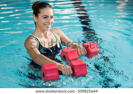 Fit woman working out with foam dumbbell in swimming pool. Woman engaged in doing aqua aerobics in water. Young beautiful woman doing aqua gym exercise with water dumbbell in swimming pool.