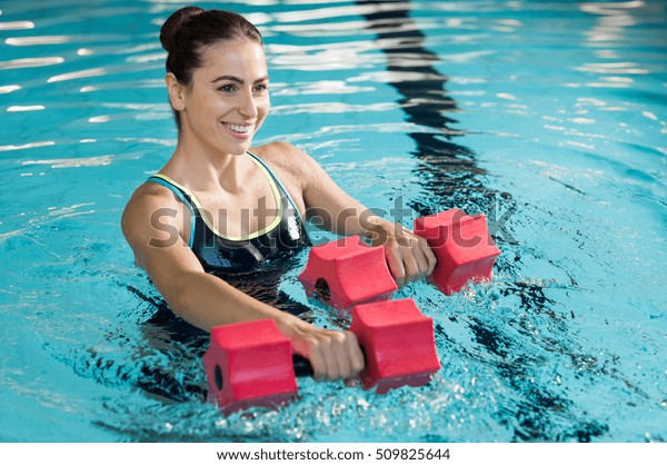 Fit woman working out with foam dumbbell in\
swimming pool. Woman engaged in doing aqua aerobics in water. Young\
beautiful woman doing aqua gym exercise with water dumbbell in\
swimming pool.