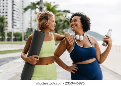 Fit women in sports clothing smile and laugh together, holding a yoga mat and a water bottle on a beach promenade. They enjoy each other's company for some outdoor exercise and a healthy lifestyle. - Powered by Shutterstock