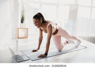 Fit young African American woman with earphones doing strength workout in front of laptop at home. Athletic black lady having fitness class on internet, watching online sports video indoors - Powered by Shutterstock