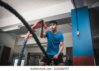 A fit young asian man working out vigorously with battle ropes. Alternating single arm waves. Whole body workout, conditioning and cardio at the gym. - Shutterstock ID 2132282797