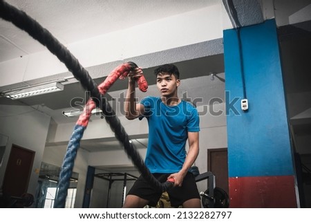 A fit young asian man working out vigorously with battle ropes. Alternating single arm waves. Whole body workout, conditioning and cardio at the gym.