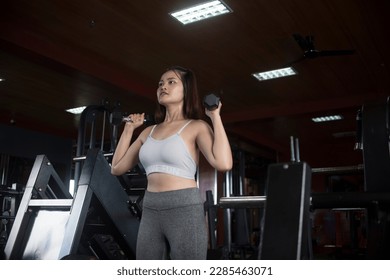 A fit young asian woman does dumbbell squats or hammer presses, holding the dumbbells in the front rack position. Training legs at a modern gym.