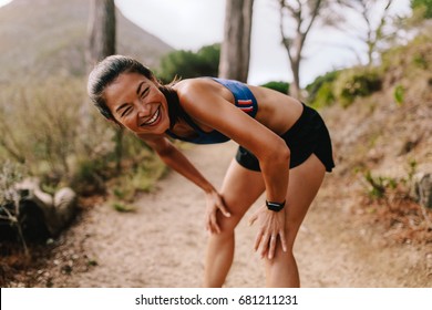 Fit young asian woman standing on mountain trail with her hands on knees and smiling. Female runner in sportswear taking a break after running workout. - Powered by Shutterstock