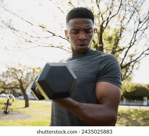 Fit young black man in t-shirt and wireless earphones doing bicep curl with dumbbell in park