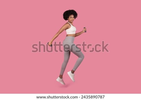 Fit young black woman with natural curly hair confidently steps forward or running while holding blue dumbbells, dressed in workout gear against pink background