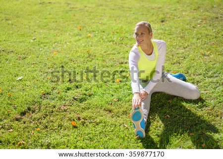 Fit young blond woman doing exercises in a park sitting on the lush green grass stretching to touch her toes, high angle view with copy space