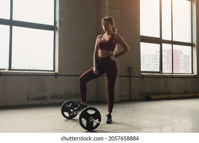 Fit Young Confident Female Athlete In Active Wear Standing On Floor With Barbell While Looking At Camera In Gym