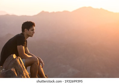Fit young man sitting alone on top of a mountain at beautiful sunset. 