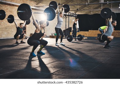 Fit young people lifting barbells over their heads looking focused, working out in a gym with other people