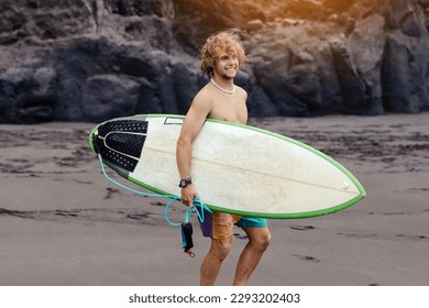 Fit young surfer man with curly blond hair with surfboard goes by the ocean having fun doing extreme water sports, surfing. Travel and healthy lifestyle concept. Sports travel destination - Powered by Shutterstock