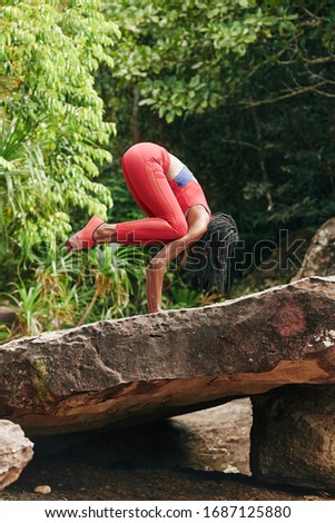 Fit young woman in bright sportswear practicing handstand on big rock in rainforest