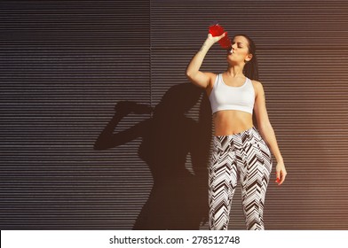 Fit Young Woman Drinking From Her Water Bottle While Standing On Black Wall Background, Female Runner In Workout Clothes Refreshing With Energy Drink During Exercise Outdoors In City At Sunset, Filter