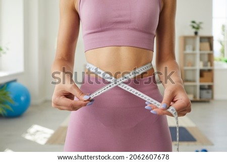 Fit young woman holding measuring tape on her thin waist. Unrecognizable slim lady in crop top showing her weightloss achievement after home workouts. Close up, closeup. Beauty and weight loss concept