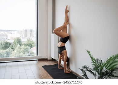 Fit young woman practices an advanced yoga pose handstand near large window overlooking city. Sportswoman in athletic wear demonstrates balance and strength in well-lit, spacious room. - Powered by Shutterstock