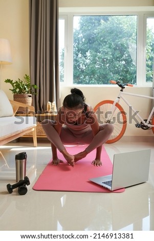 Fit young woman practicing handstand on yoga mat following tutorial on laptop at home