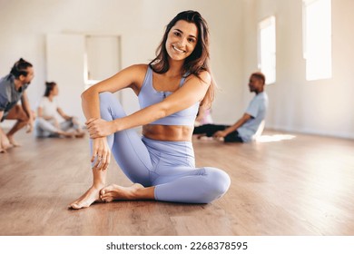 Fit young woman smiling at the camera while sitting in a yoga studio with her class in the background. Happy young woman having a workout session with her class in a fitness studio. - Powered by Shutterstock